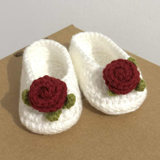 White crocheted slippers with red rose decorations on a beige surface.